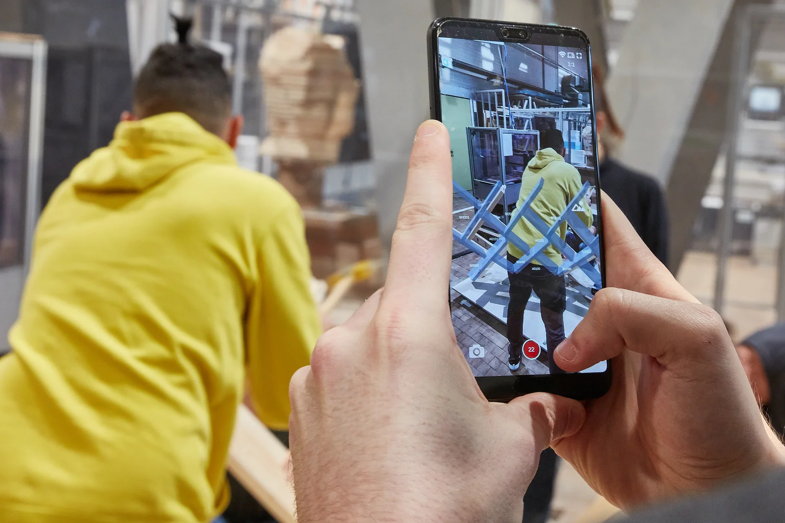 Timber construction with the help of virtual reality Blick auf ein Smartphone Display, welches eine Aufbauanleitung für einen Holzaufbau anzeigt. Diese Anleitung ist durchscheinend, so dass auf dem Display ebenfalls eine Person sichtbar ist, welche ein nummeriertes Bauteil an der richtigen Stelle einbaut. Es handelt sich also um eine Art virtuelle Aufbauanleitung für ein Holzständerbauwerk.