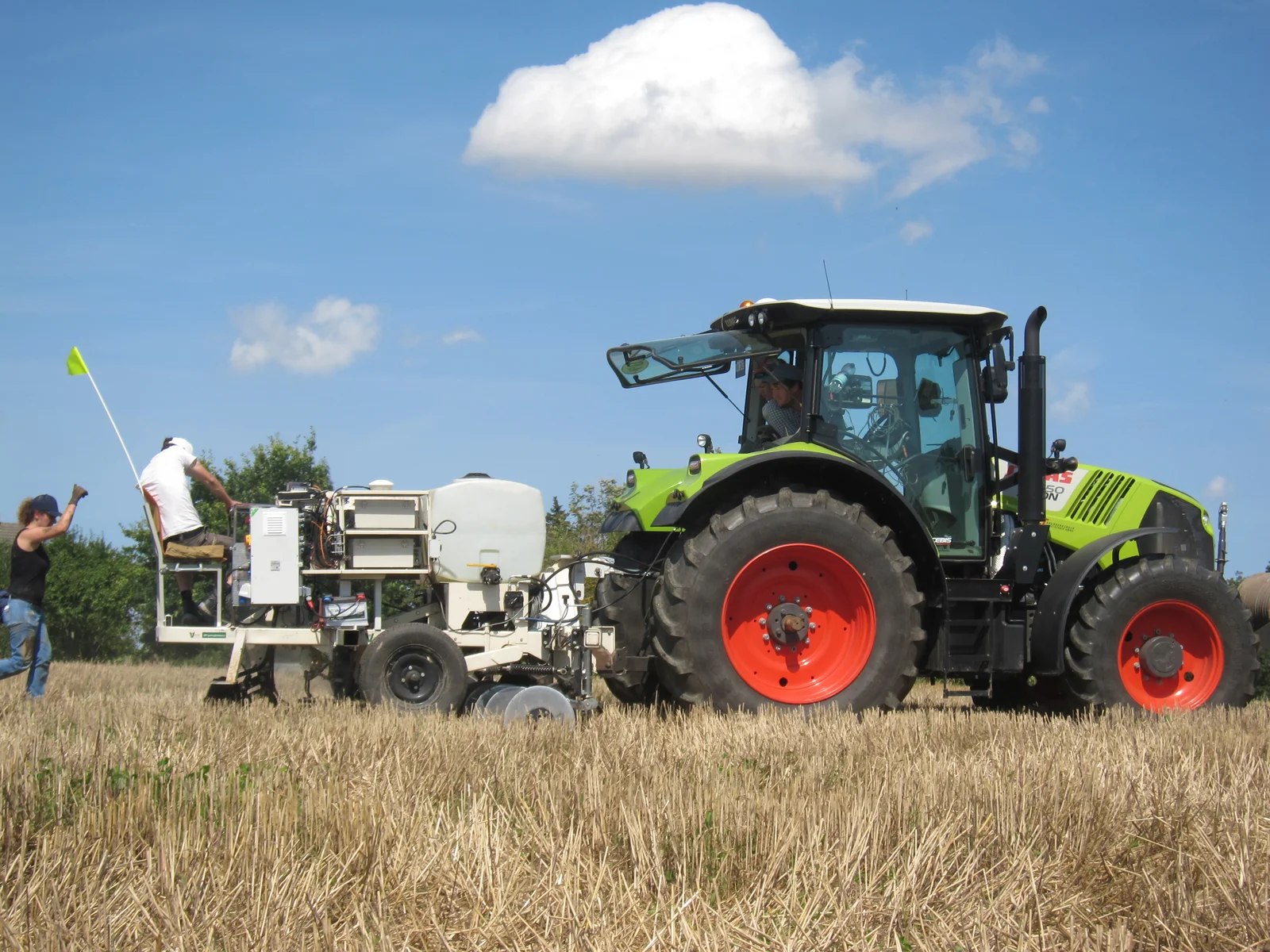 Tractor pulls measurement technology Ein Traktor zieht eine Vorrichtung zur präzisen Messung des ph-Wertes über einen Stoppelacker. Auf der Messvorrichtung sitzt ein Wissenschaftler und schaut auf den Ackerboden. Hinter der Messvorrichtung läuft eine Wissenschaftlerin. Sie schaut ebenfalls auf den Ackerboden und gibt mit dem Daumen nach oben ein positives Signal an den Wissenschaftler und den Traktorfahrer.