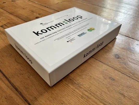 Planspiel komm:loop Eine Pappverpackung des Planspiels liegt auf einem Holztisch.
