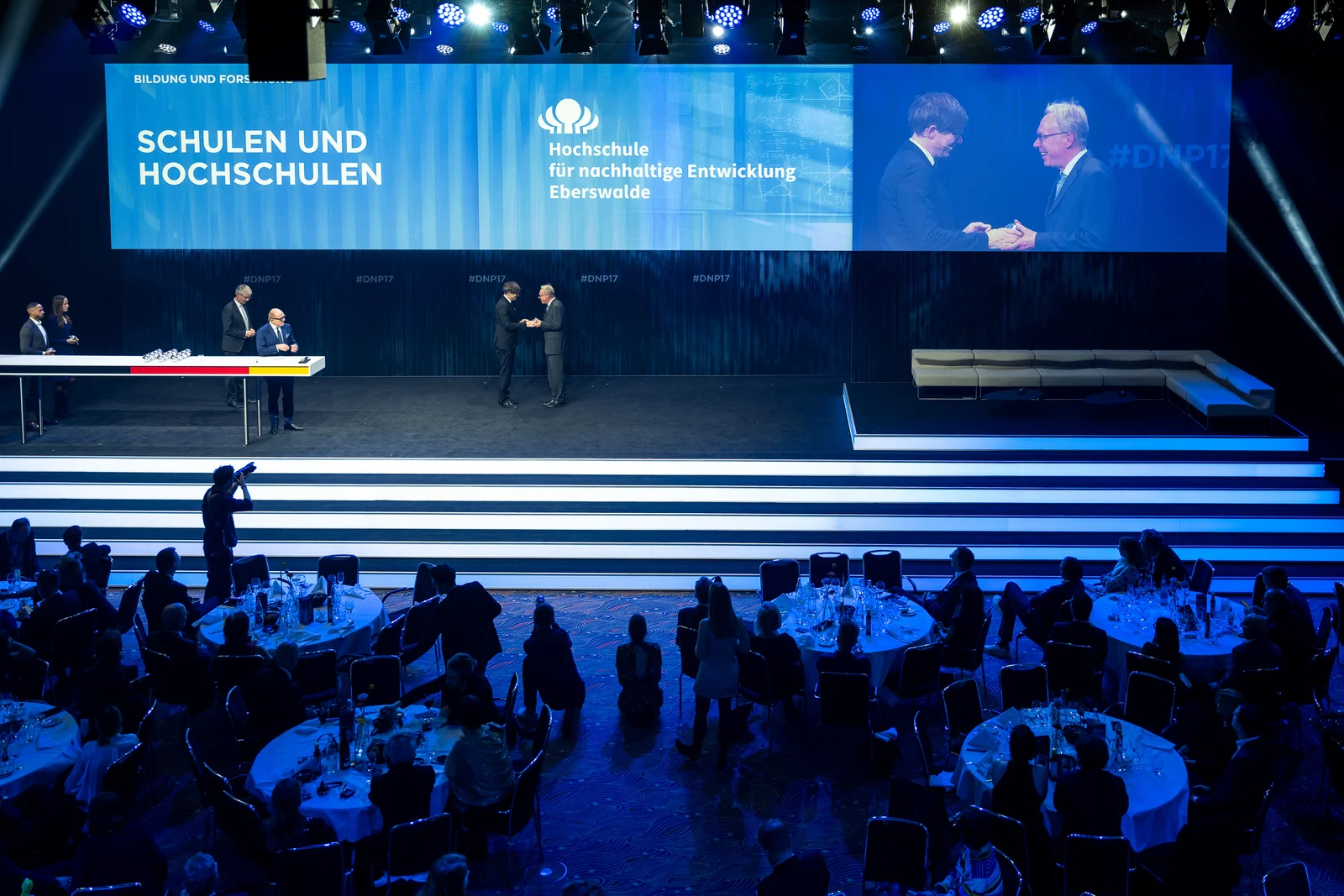 Zwei Männer schütteln sich auf der Bühne des Deutschen Nachhaltigkeitspreises 2024 die Hände. An die Wand sind Logo und Name der Hochschule für nachhaltige Entwicklung Eberswalde projiziert. Links sieht man die Kategorie Bildung und Forschung und darunter Schulen und Hochschulen