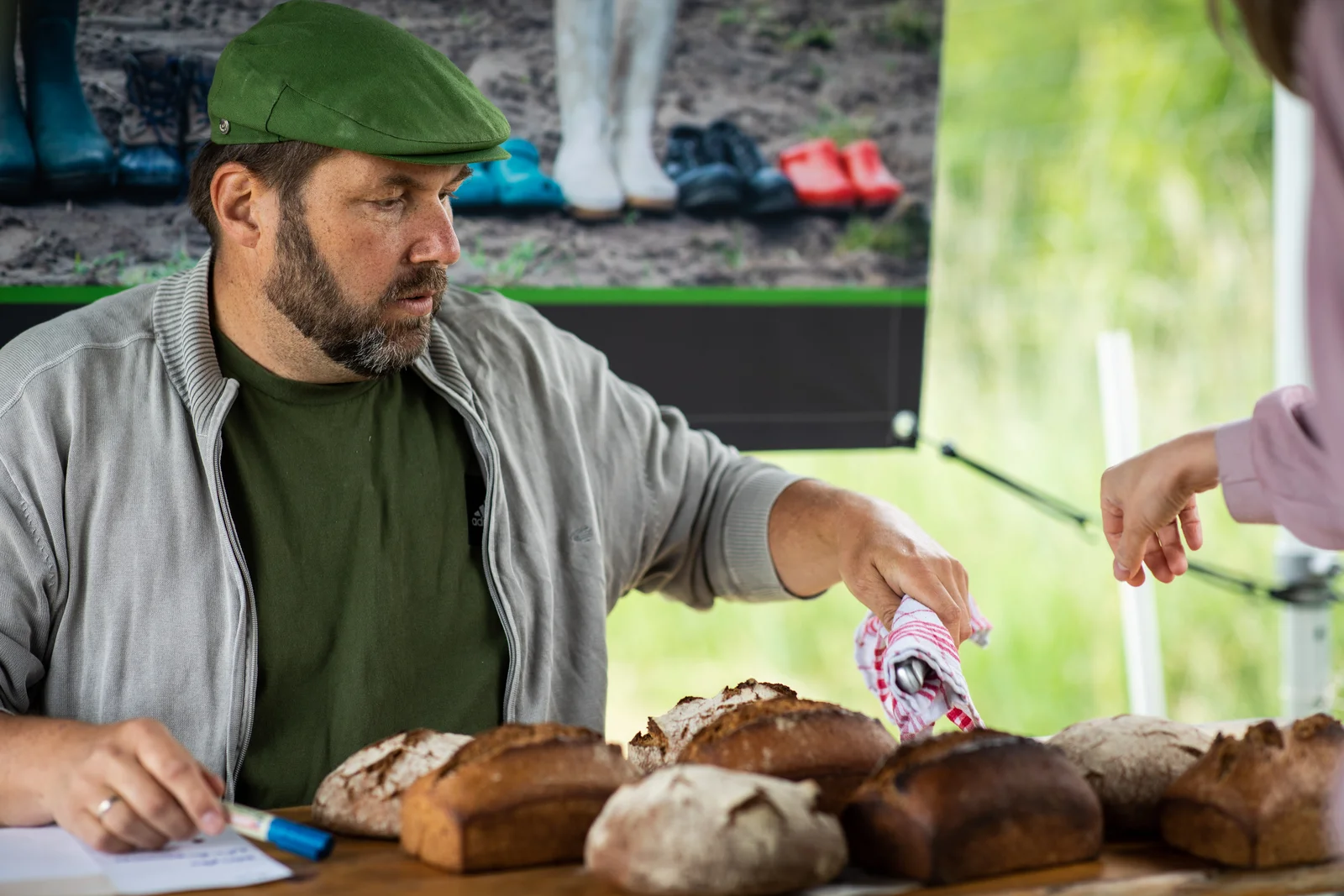 Bäcker Björn Wiese beim Feldtag Ein Mann sitzt an einer Biertischgarnitur. Hinter ihm ist ein Banner mit abgebildeten Gummistiefeln zu sehen. Im Hintergrund erkennt man, dass es eine Situation in der draußen ist. Vor dem Mann liegen unterschiedliche Brote. Er hält ein Brotmesser in der Hand.