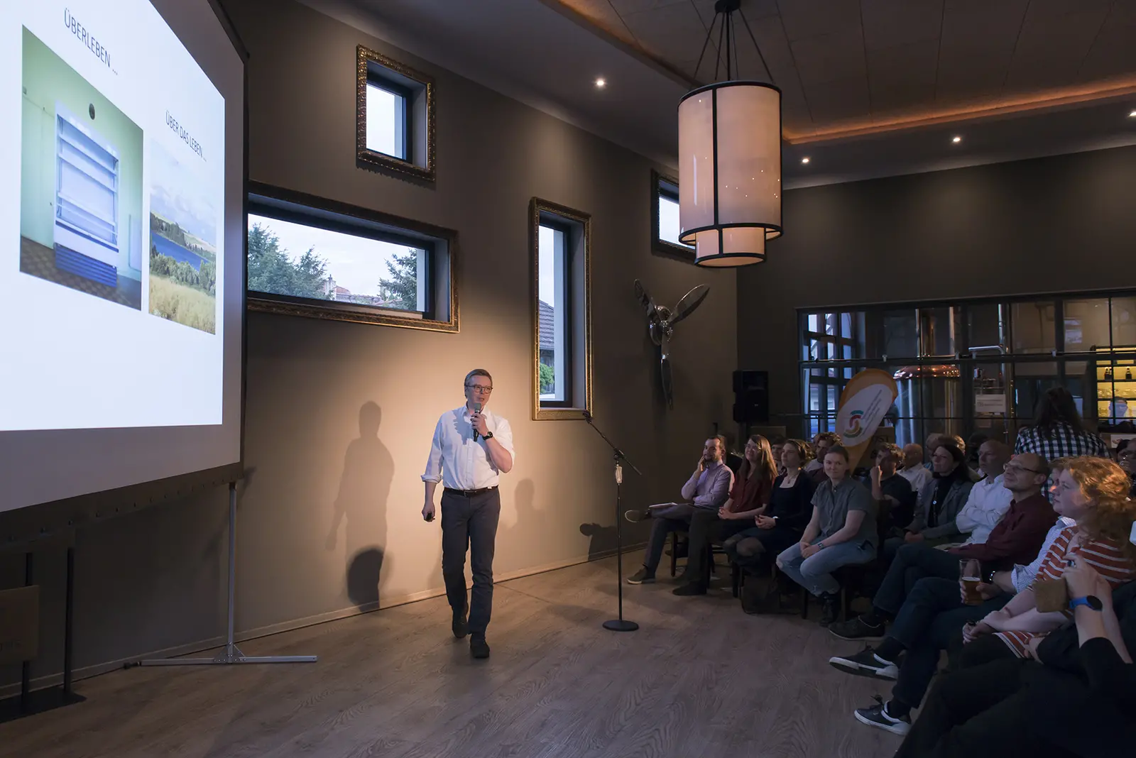 Science Slam Präsenzstelle 2022 Das Bild zeigt einen Sprecher während des Brandenburger Science Slam in Schwedt. Er steht vor einem Publikum und präsentiert auf einer Leinwand Bilder von zwei unterschiedlichen Landschaften. Die Zuhörer sitzen auf Stühlen und hören aufmerksam zu.