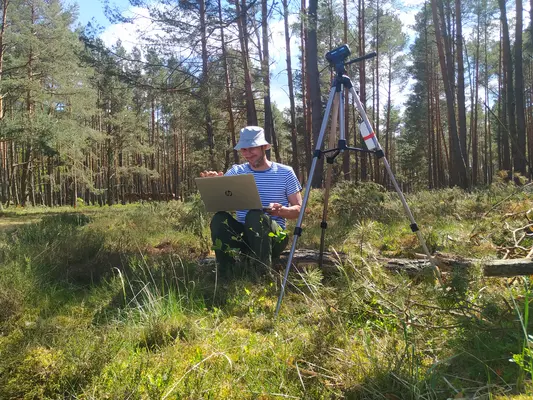 Dr. Jakob Wegener, wissenschaftlicher Mitarbeiter der HNEE und Gründer der GrüBie Gbr Abgebildet ist ein Kiefernwald mit moosiger Vegetation im Vordergrund. Mittig sitzt Dr. Jakob Wegener auf einem Baumstamm. Er blickt auf seinen aufgeklappten Laptop auf seinem Schoß. Auf dem Bild rechts von ihm steht eine Kamera für die Beobachtung der Bienenvölker.
