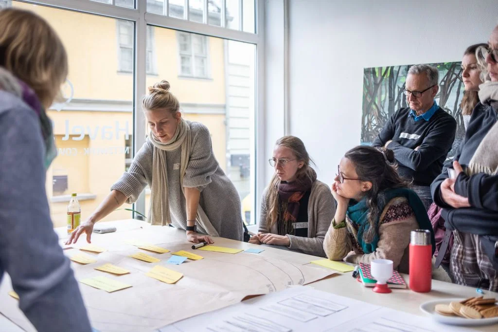 Workshop mit Projektpartner*innen Um einen Tisch stehen und sitzen mehrere Menschen. Auf dem Tisch zwischen ihnen liegen große Flipchartpapiere. Eine Frau erläutert gerade den Inhalt eines dieses Papiers.