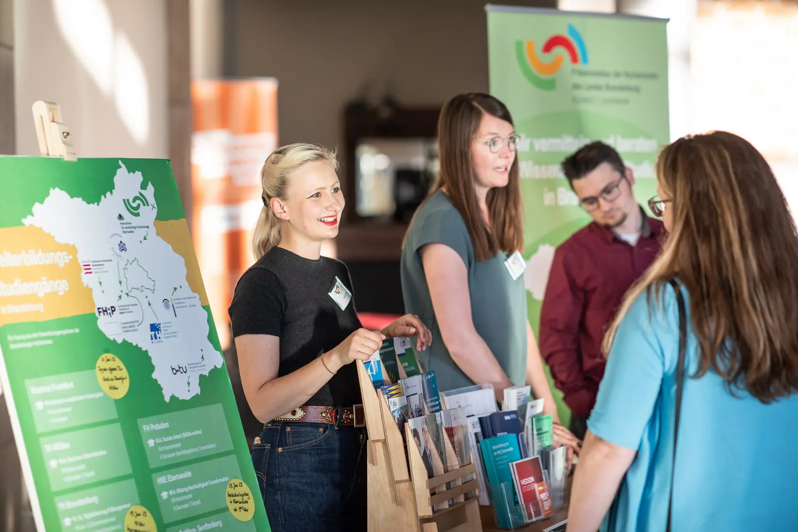 Präsenzstelle Schwedt | Uckermark Auf dem Bild ist eine Frau zu sehen, die lächelnd an einem Informationsstand steht. Sie hält Informationsmaterial in der Hand und unterhält sich mit einer Person, die nicht im Bild zu sehen ist. Im Hintergrund befindet sich ein Poster mit einer Landkarte und Informationen über Studienmöglichkeiten in Brandenburg.
