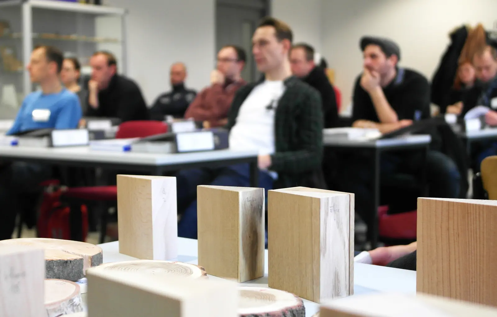 Studierende in einer Vorlesung im Vordergrund stehen Holzblöcke im Hintergrund sieht man verschwommene Menschen sitzen.