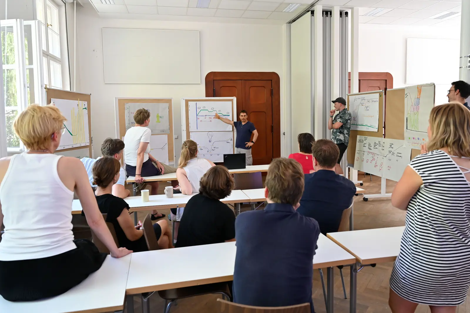 Workshop im Seminarraum Ein Gruppe diskutiert in einem Seminarraum. Mehrere Stellwände mit während des Workshops erarbeiteten Abbildungen stehen im Halbkreis. Ein Mann erklärt gerade eine dieser Abbildungen.
