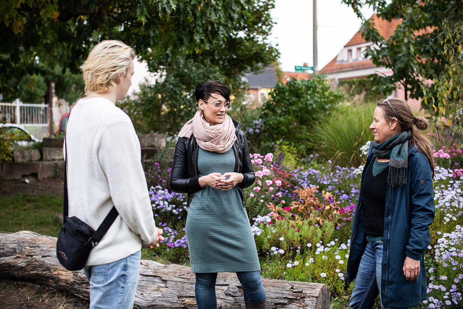 Service Learning Drei Personen stehen vor einer bunt blühenden mit Stauden bepflanzten und von Bäumen beschatteten Fläche. Im Hintergrund sieht man Häuser. Es handelt sich um den sogenannten Blumenplatz in Eberswalde. Eine Dozentin der Hochschule ist im Gespräch mit einer Partnerin der Zivilgesellschaft und einem Studierenden.