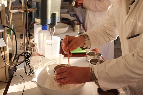 Laboratory internship Ein Forscher im Laborkittel siebt eine Bodenprobe im Labor. Er streicht mit einem Pinsel über das Sieb und die Bodenteilchen rieseln in ein Gefäß. Daneben steht ein Labormixer.