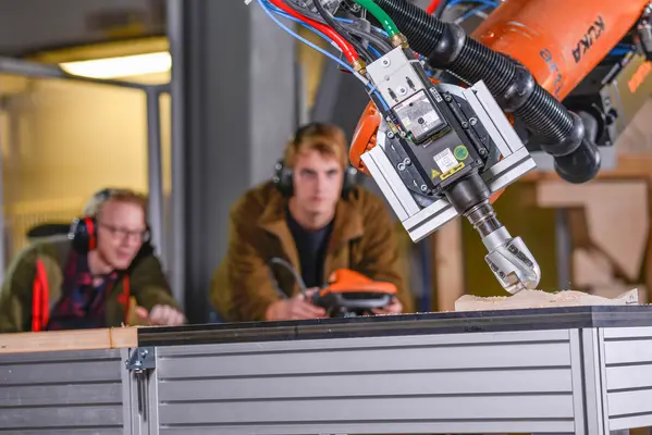 Robot arm with milling cutter Roboterarm mit eingespanntem Fräskopf bearbeitet einen Holzprobekörper. Im Hintergrund sieht man zwei junge Wissenschaftler mit der tragbaren Robotersteuerung in der Hand.