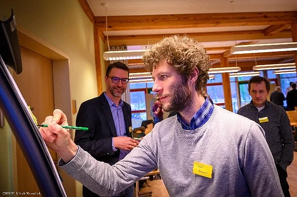 InnoForum Wald und Holz Das Bild zeigt drei Personen. Der Mann im Vordergrund schreibt etwas auf ein Whiteboard während zwei Personen im Hintergrund zusehen