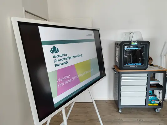 3D-Print Workshop Im Vordergrund ist ein digitales Board mit Begrüßung abgebildet. Im Hintergrund steht der Flashforge 3-D-Drucker.