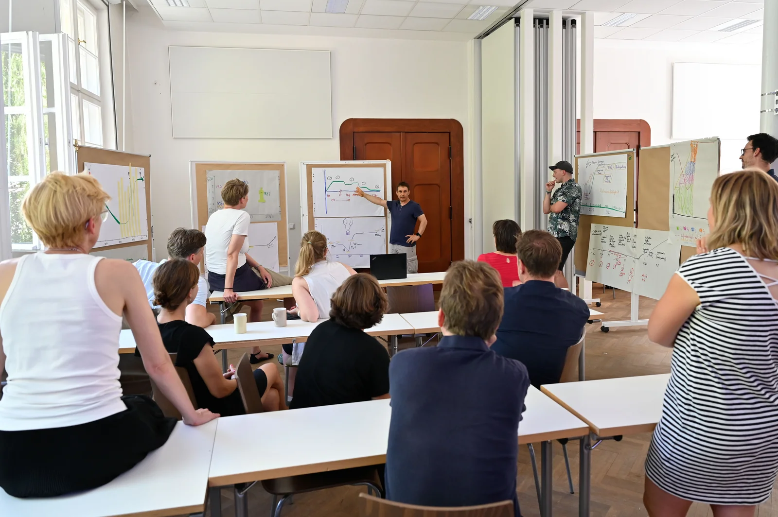 Workshop in the seminar room Ein Gruppe diskutiert in einem Seminarraum. Mehrere Stellwände mit während des Workshops erarbeiteten Abbildungen stehen im Halbkreis. Ein Mann erklärt gerade eine dieser Abbildungen.